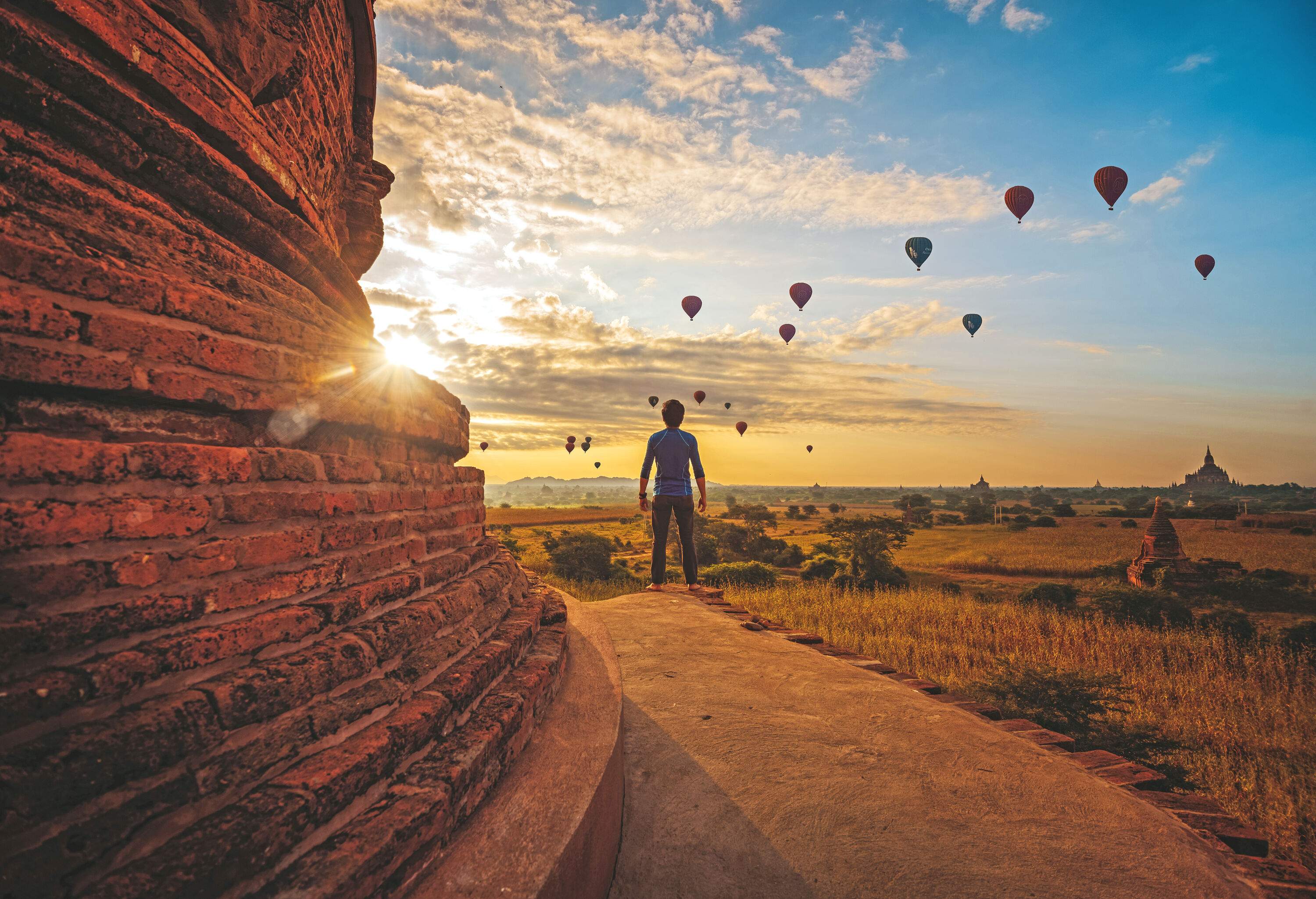 A man stands beside a ruined temple wall, watching the hot air balloons flying against the scenic sky.