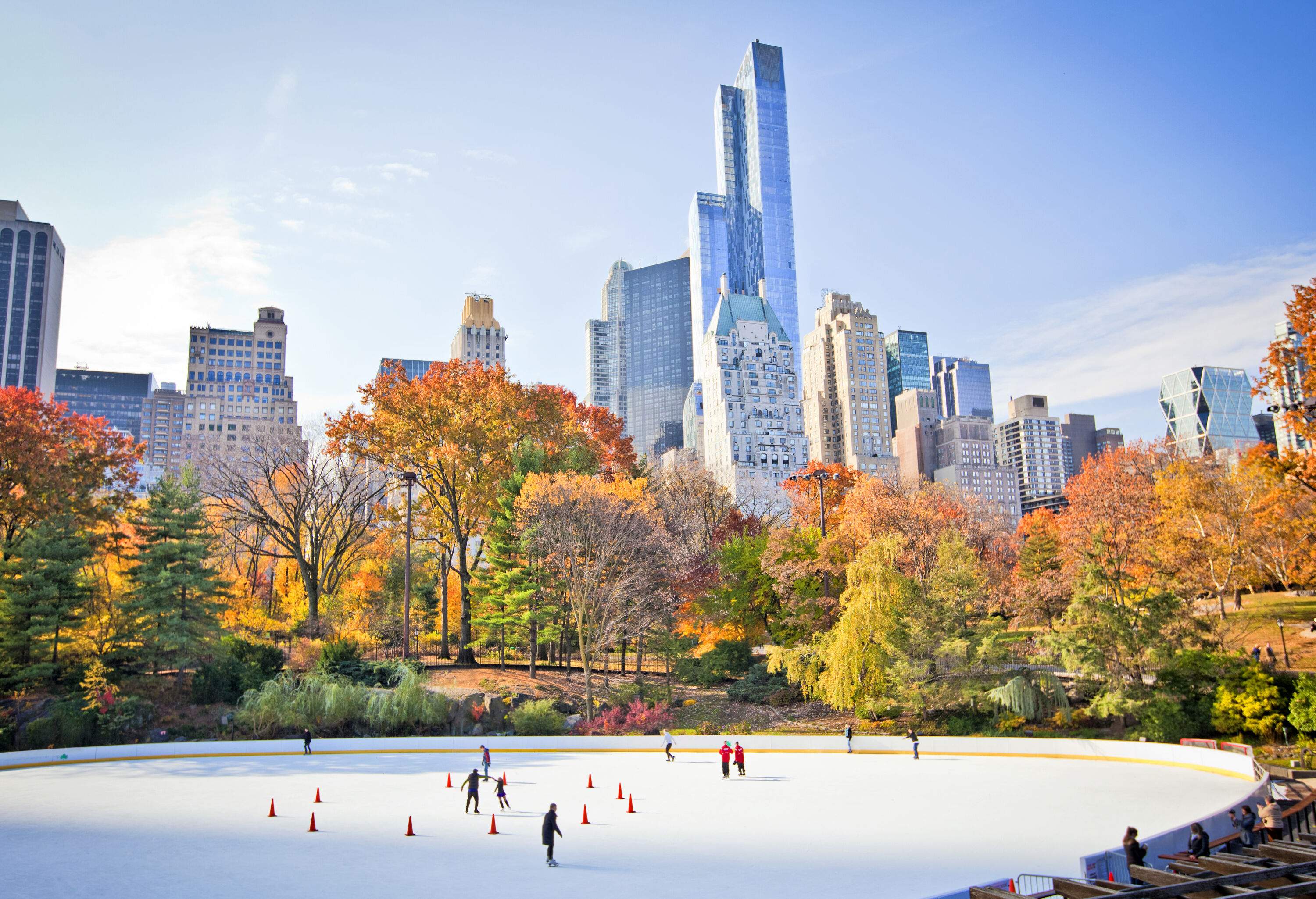 People skating on a park's ice rink surrounded by colourful autumn trees with views of modern skyscrapers.