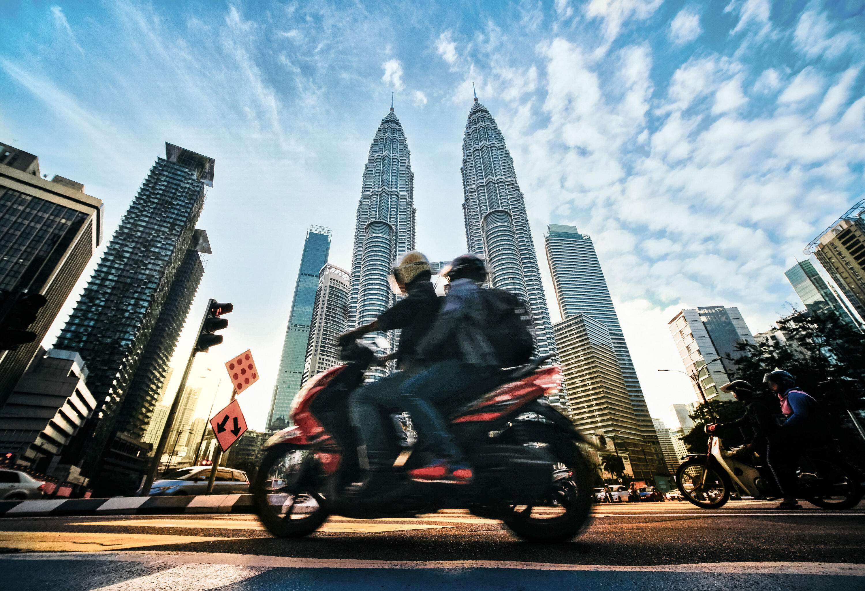 Motorists on a bustling street along the majestic Petronas Twin Towers between tall buildings.