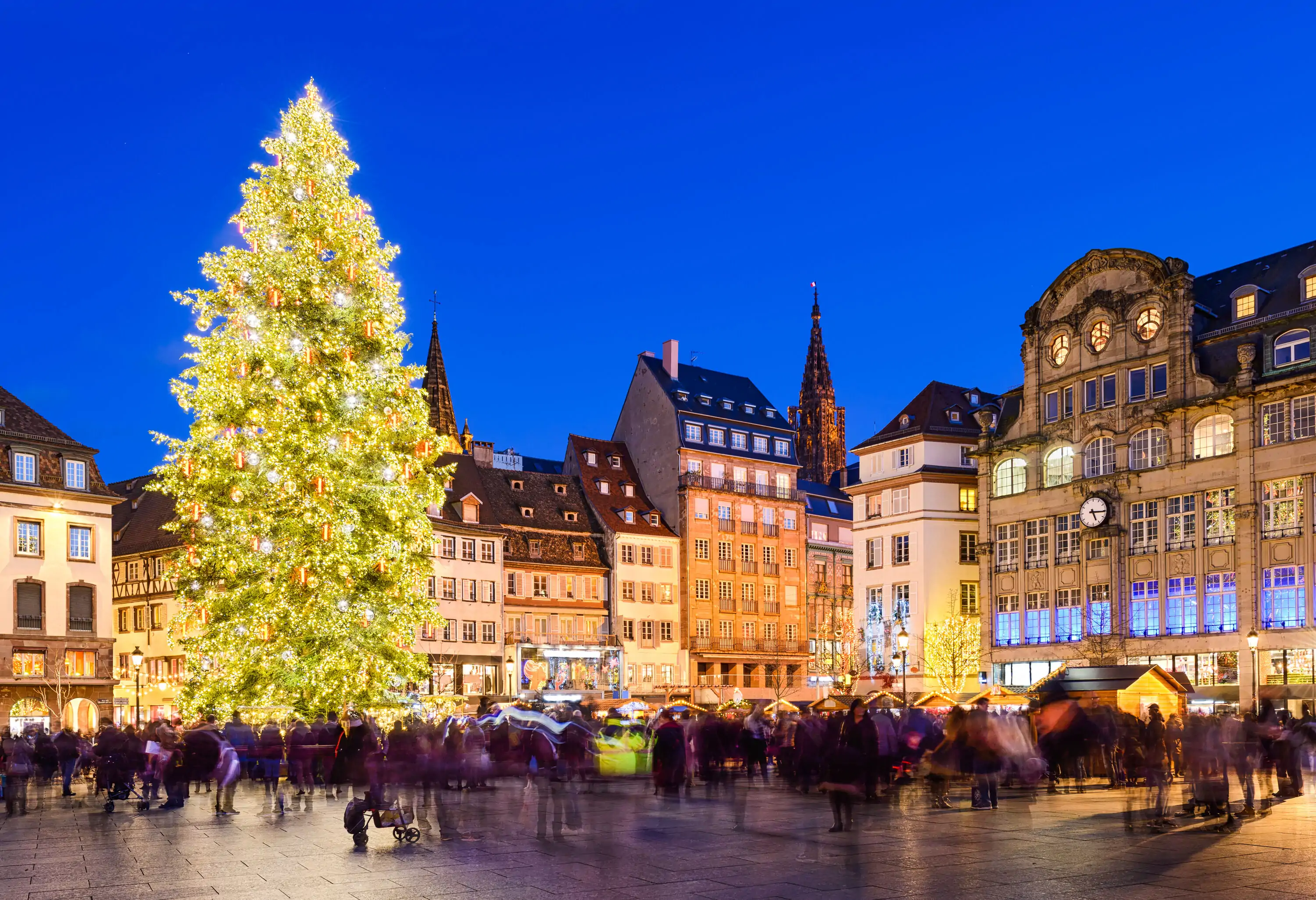 A bright and dazzling Christmas tree towering over a plaza filled with people.