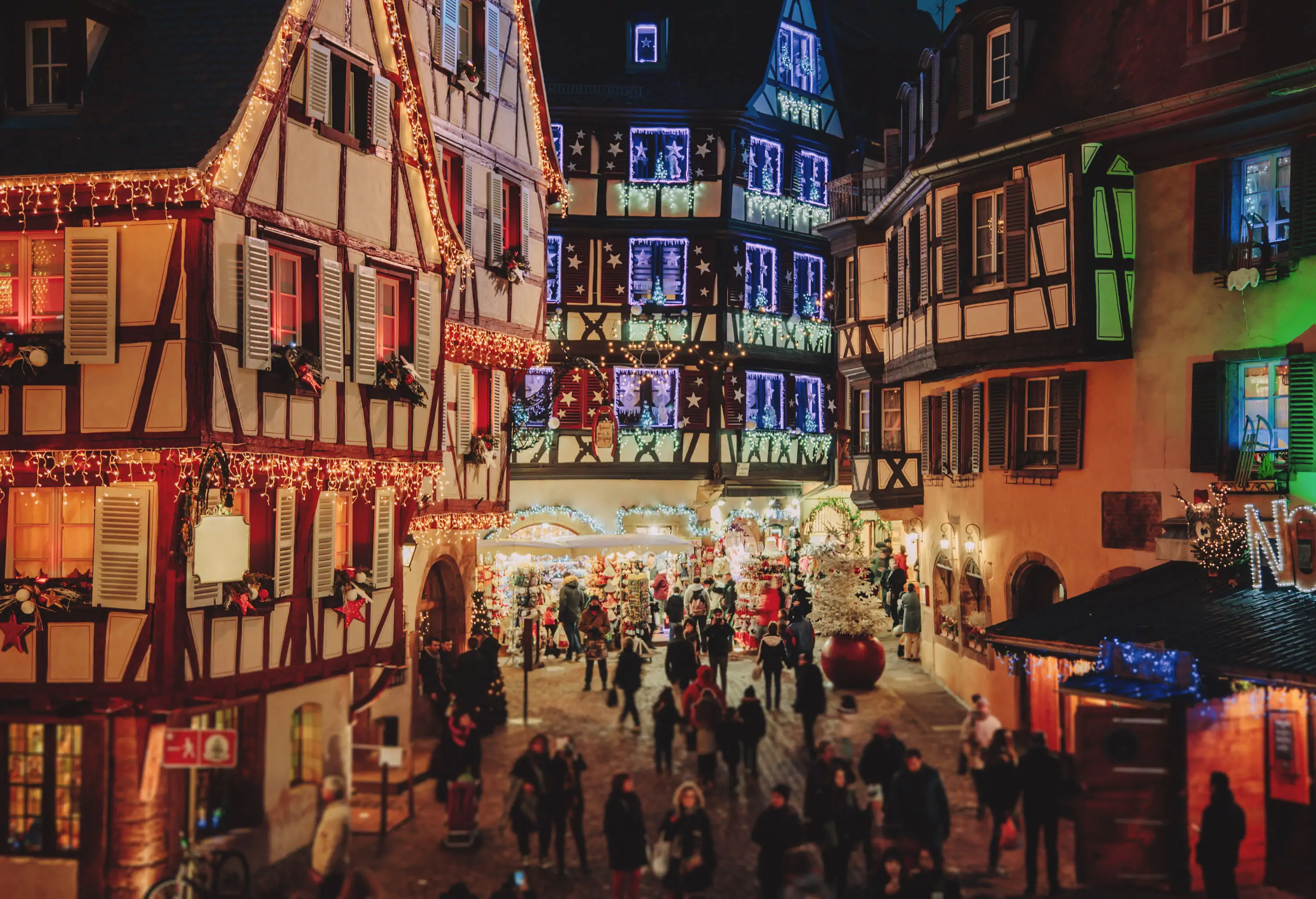 People are in the street surrounded by traditional half-timbered houses decorated with colourful lights and Christmas ornaments.