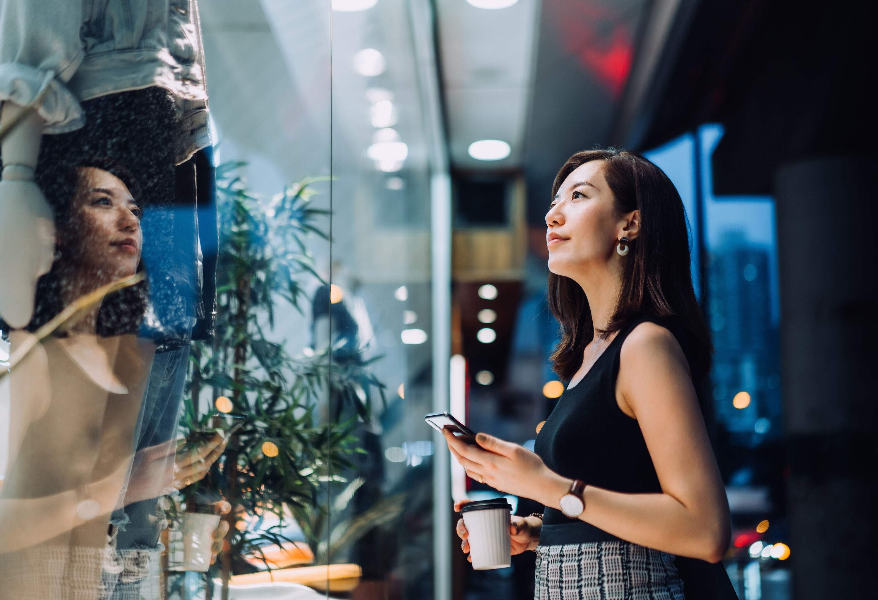 A classy woman with a coffee cup and a smartphone looking at the clothes displayed in a store window.