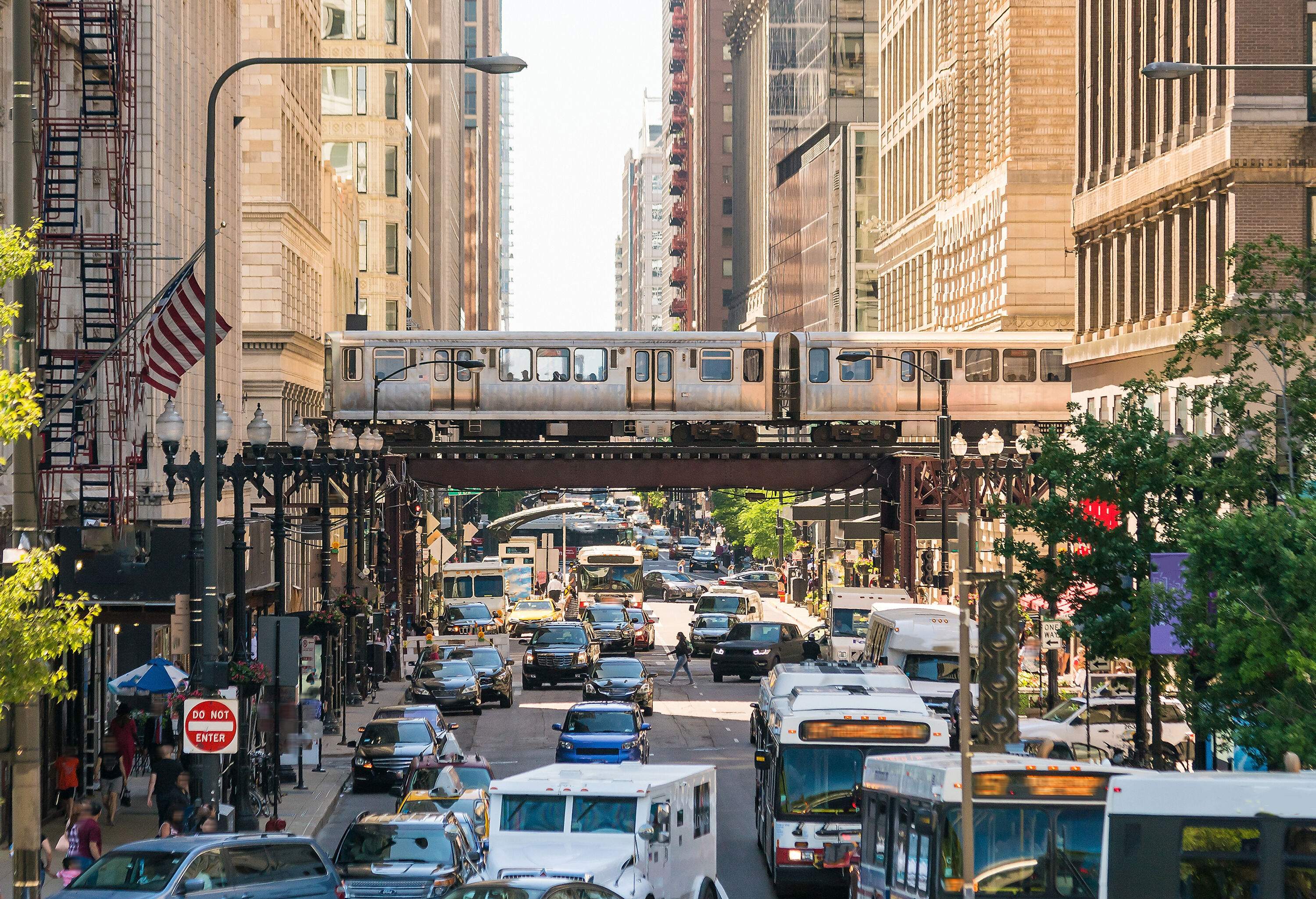 A grey train travelling on an elevated railway across the busy street between the tall buildings.