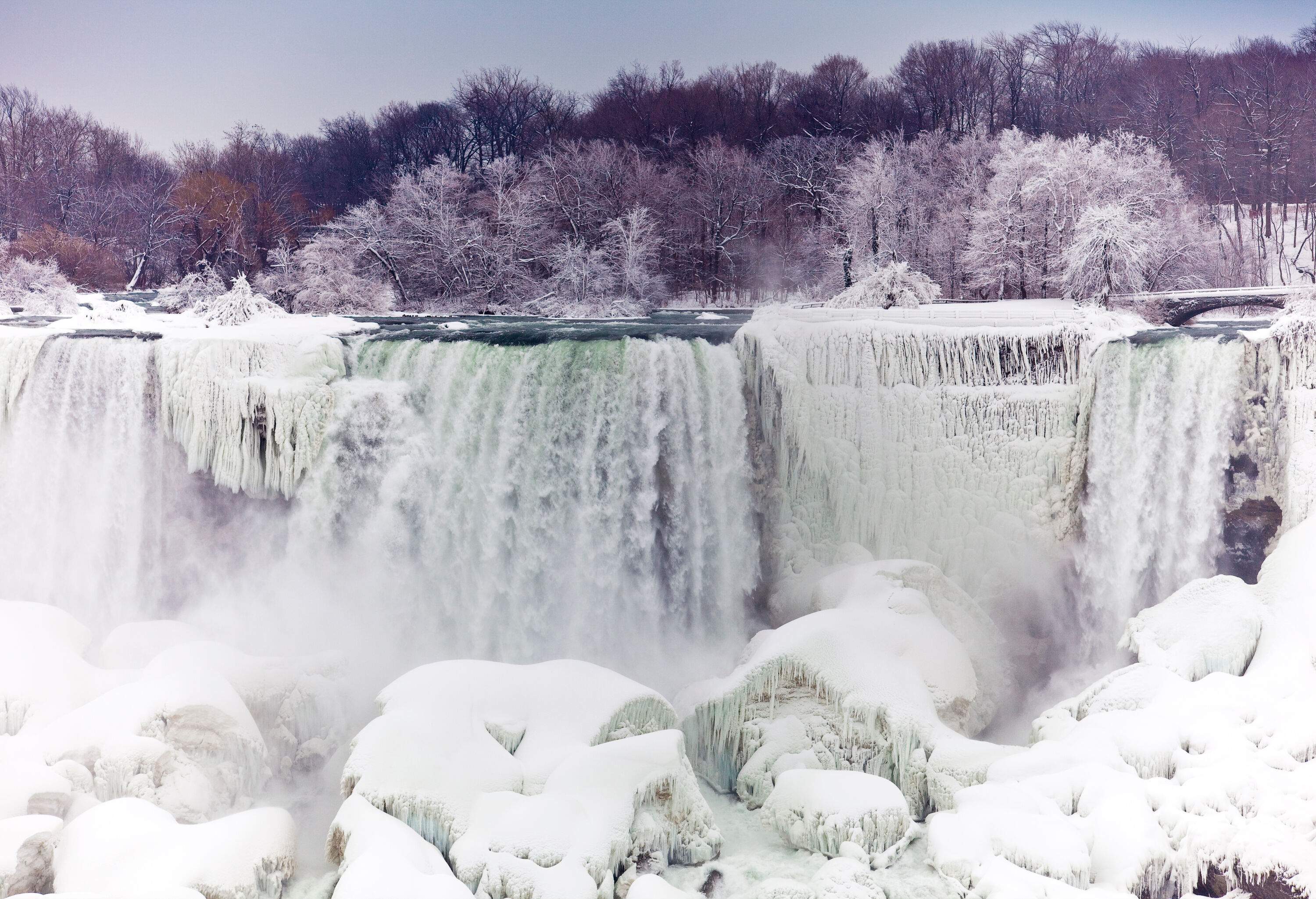 A frozen waterfall surrounded by frosted trees and snow-covered boulders.