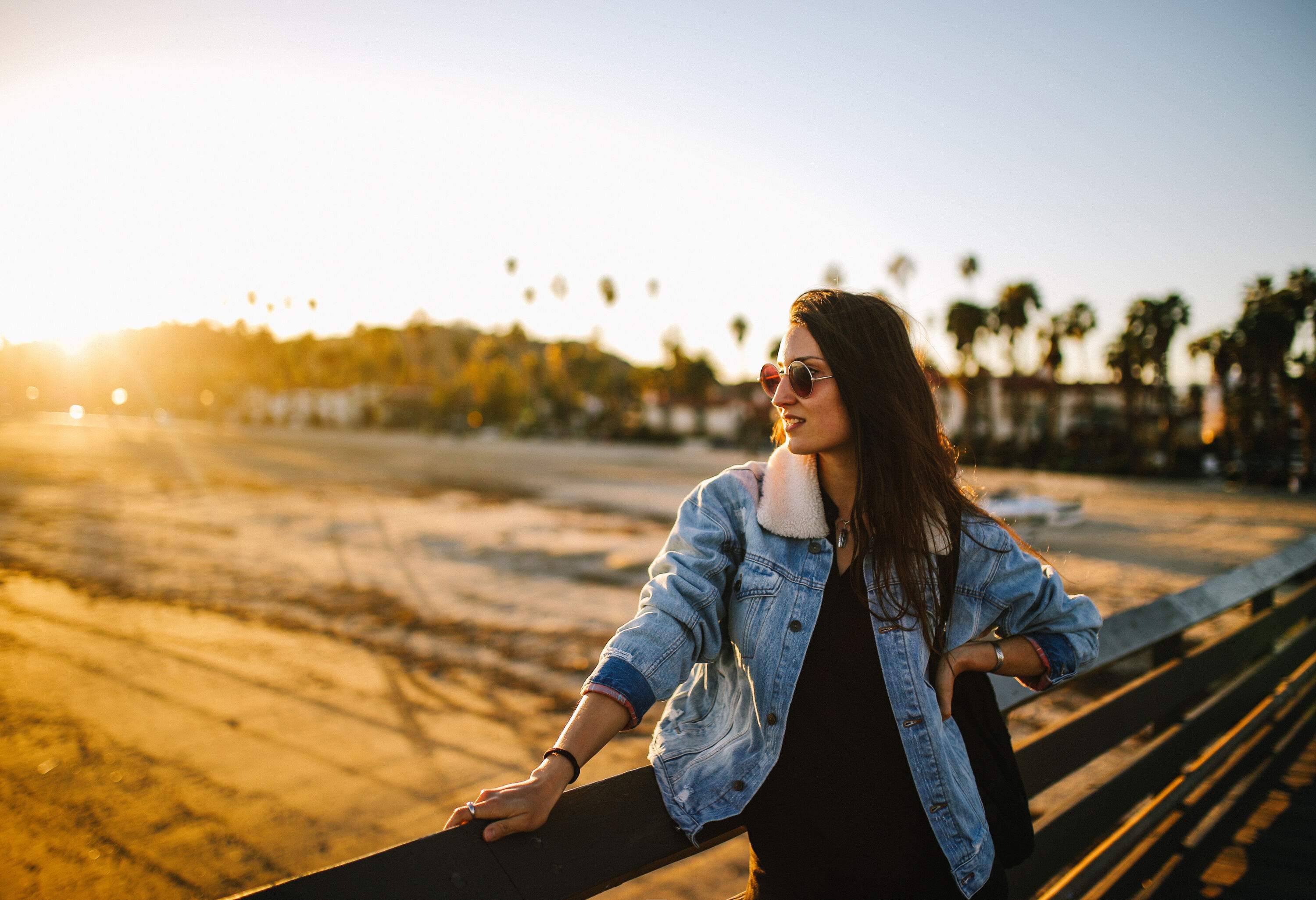 A long-haired lady in a denim jacket wears sunglasses against the sun's rays while leaning on a wooden fence at a boardwalk.